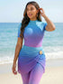 A woman stands on a sandy beach wearing a fitted short-sleeve top and matching leggings with a blue-to-purple gradient color shift, plus a matching draped wrap skirt with a gold flower detail.