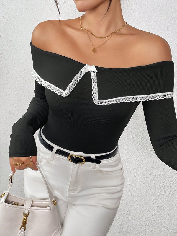 A woman wearing a black off-the-shoulder long-sleeve top with white lace trim, paired with white pants and a black belt, holding a white handbag against a textured white wall.