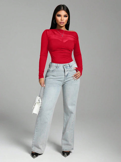 A woman modeling a bright red long-sleeve top with sheer mesh panels and light-wash wide-leg jeans, standing against a plain gray studio background.