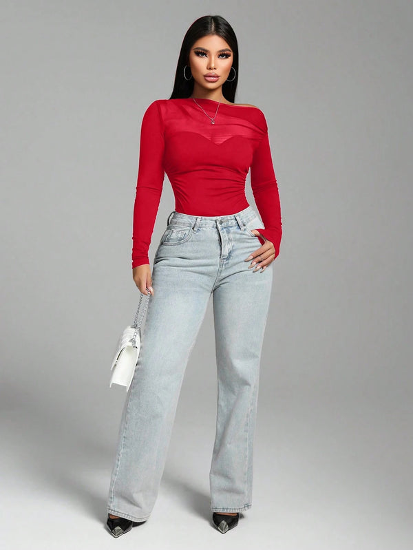 A woman modeling a bright red long-sleeve top with sheer mesh panels and light-wash wide-leg jeans, standing against a plain gray studio background.