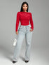 A woman modeling a bright red long-sleeve top with sheer mesh panels and light-wash wide-leg jeans, standing against a plain gray studio background.