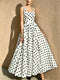 A woman models a sleeveless white A-line maxi dress with a black polka dot pattern, standing against a textured beige wall.