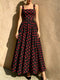 A woman models a sleeveless black maxi dress with a red polka dot pattern, standing against a textured beige stucco wall.
