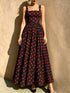 A woman models a sleeveless black maxi dress with a red polka dot pattern, standing against a textured beige stucco wall.