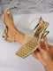 A pair of metallic gold high-heeled sandals with a mesh upper and ankle straps, shown on a foot and held by a hand against a light gray background with draped fabric and a pearl necklace.