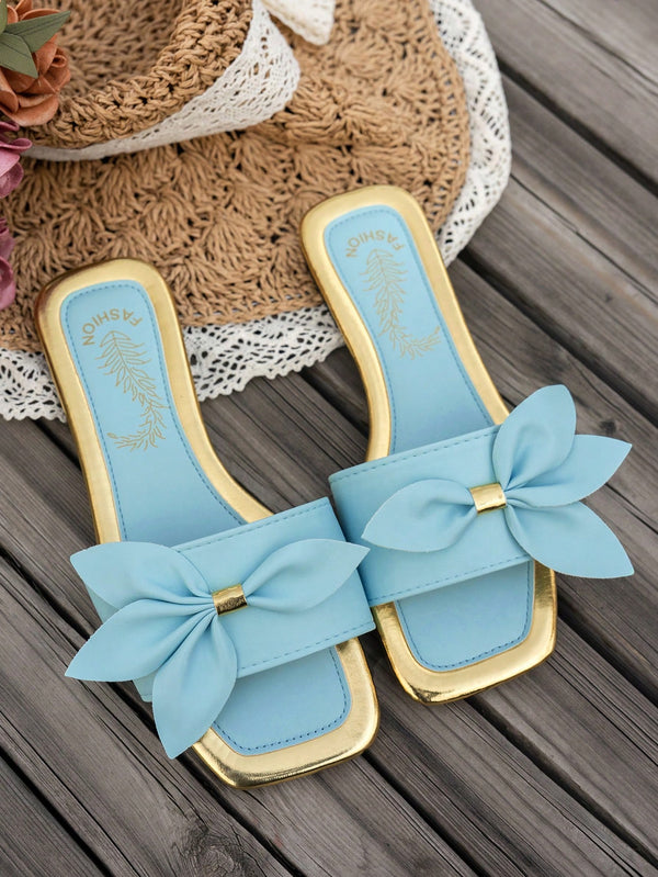 A pair of light blue slide sandals with gold trim and large fabric bow details on the toes, displayed on a dark wooden surface with a straw hat and lace mat as props.
