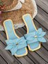 A pair of light blue slide sandals with gold trim and large fabric bow details on the toes, displayed on a dark wooden surface with a straw hat and lace mat as props.