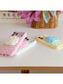 Three decorative phone cases with 3D rose designs are displayed on a tabletop. One pink case with white polka dots and a white rose is in the foreground, a white case with pink polka dots and a pink rose is behind it, and a pale yellow case with blue polka dots and a blue rose is on the right.