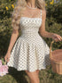 A woman wearing a white sleeveless mini dress with black polka dots and thin shoulder straps, standing outdoors in a grassy area with greenery and holding a wicker basket and pink flowers.