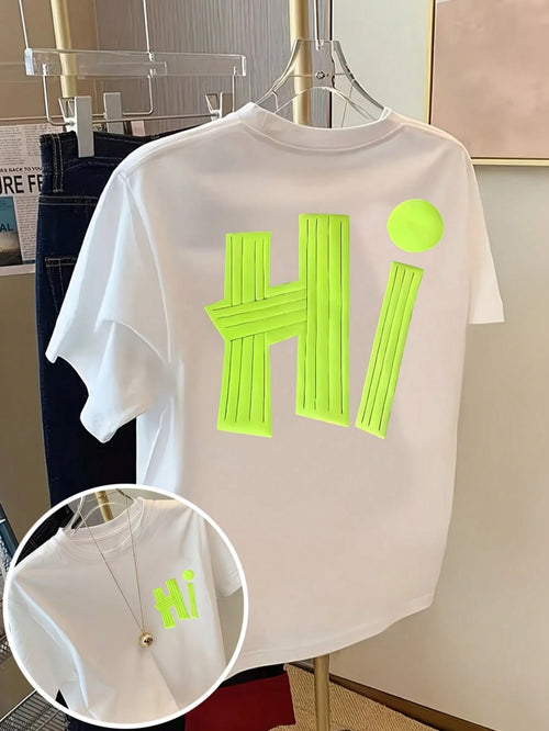 A white short-sleeve T-shirt hanging on a clear hanger, featuring a large neon yellow-green 3D block letter 'Hi' on the back with a matching neon yellow-green dot above it. A circular inset shows a close-up of the neon graphic on the front chest with a gold necklace worn over it.