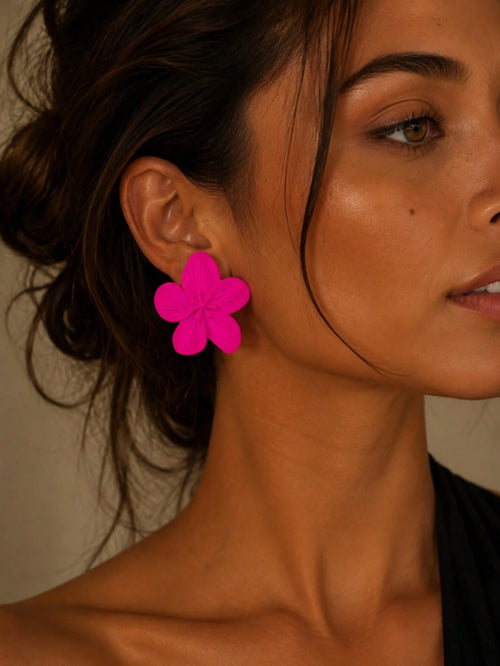 A close-up of a woman wearing a bright hot pink flower-shaped dangle earring. The earring has five layered petals and is shown on her earlobe against a warm, neutral background.
