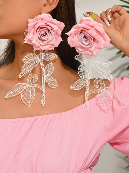 A close-up of a person wearing large pink rose earrings with white lace vine and leaf details, paired with a pink off-the-shoulder top.