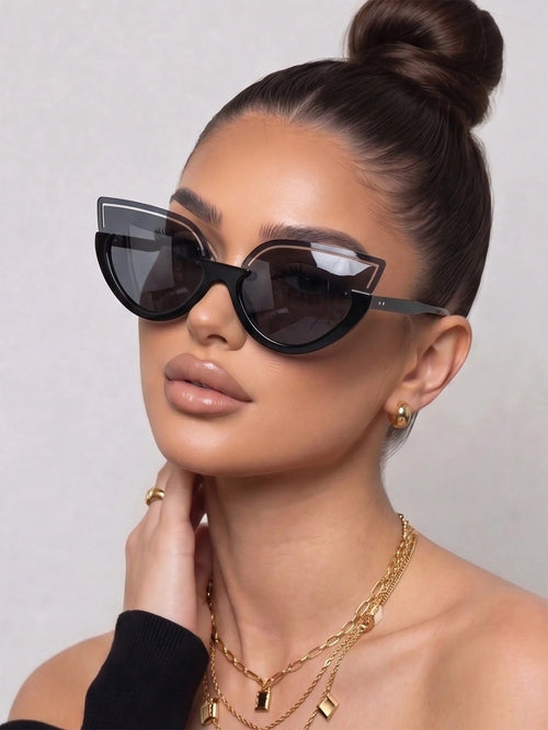 A close-up of a model wearing black cat-eye sunglasses with dark lenses, styled with gold necklaces and earrings against a neutral light gray background.