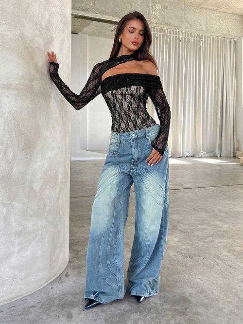 A woman modeling a black sheer lace off-the-shoulder long-sleeve top paired with wide-leg light blue jeans, standing in a modern studio with concrete floors and draped white curtains.
