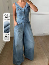 A woman wearing a light blue denim wide-leg jumpsuit in an indoor setting with a wood floor and white wall. A vertical graphic on the left highlights the cotton fabric, showing a close-up of the denim texture and icons for breathability, comfort, and lightweight.
