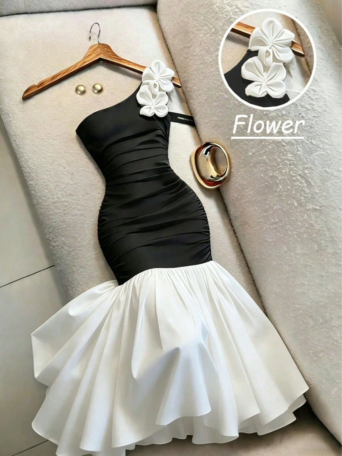 A one-shoulder black and white mermaid-style dress with white fabric flowers on the shoulder, displayed on a wooden hanger on a beige textured sofa. A circular inset shows a close-up of the white flower detail, with the word 'Flower' written next to it.