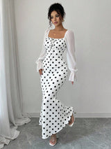 A woman models a white maxi dress with black polka dots and sheer white long sleeves, standing in a minimalist room with a light gray wall and floor.