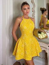 A woman models a bright yellow halter-neck mini dress with a textured floral pattern, holding a bouquet of white roses in an elegant indoor setting with a mirror reflecting her back.