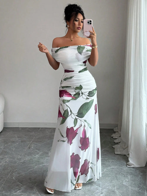A woman takes a mirror selfie wearing a white off-the-shoulder maxi dress with a large green and dark pink floral print. She is standing in a room with gray tiled floors, a light gray wall, and white curtains.