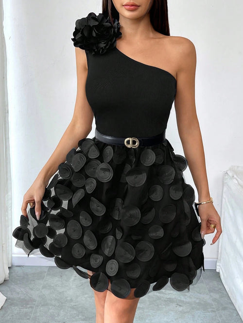 A woman wearing a black one-shoulder mini dress with a large fabric flower on the shoulder, a fitted bodice, a thin black belt with a gold-tone buckle, and a voluminous skirt covered in layered circular black fabric discs.