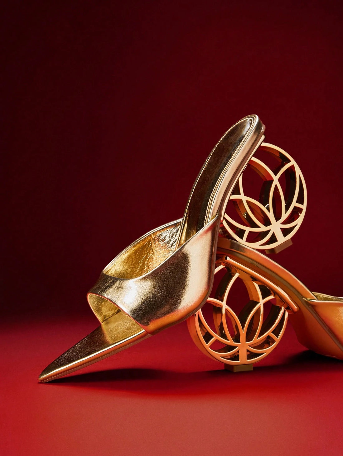 A pair of metallic gold pointed-toe mule heels with sculptural circular cutout heels, displayed on a red studio background.