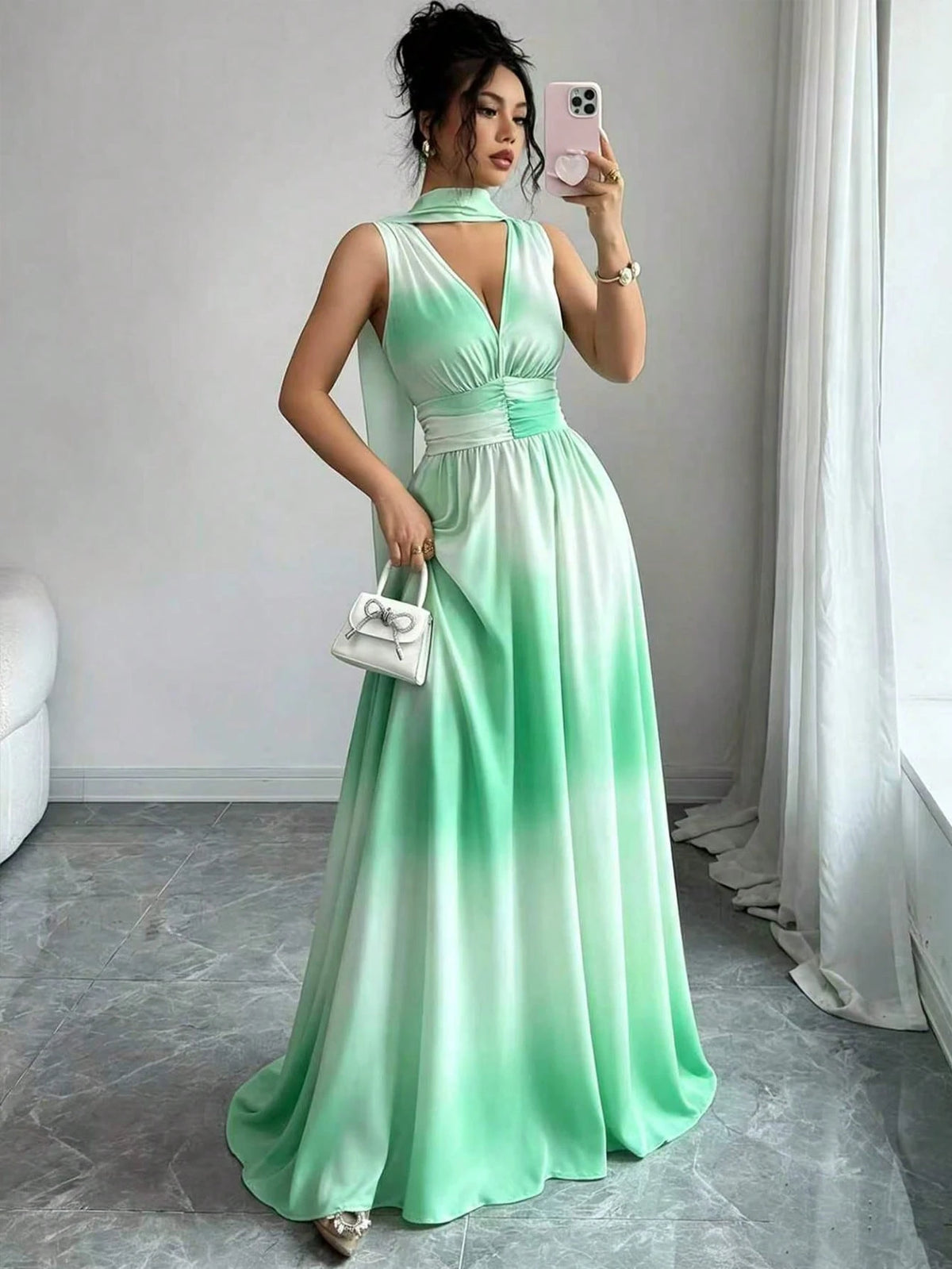A woman takes a mirror selfie wearing a sleeveless mint green to white ombré maxi dress with a halter neckline, a fitted waist, and a flowing skirt. She is holding a small white handbag in a room with gray tiled floors and white curtains.