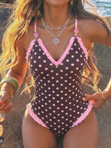 A woman wearing a one-piece swimsuit with a dark brown base and pink polka dots, featuring pink ruffled trim along the straps, neckline, and hem. The photo is taken outdoors near the water, with warm sunlight and a blurred rocky shoreline in the background.