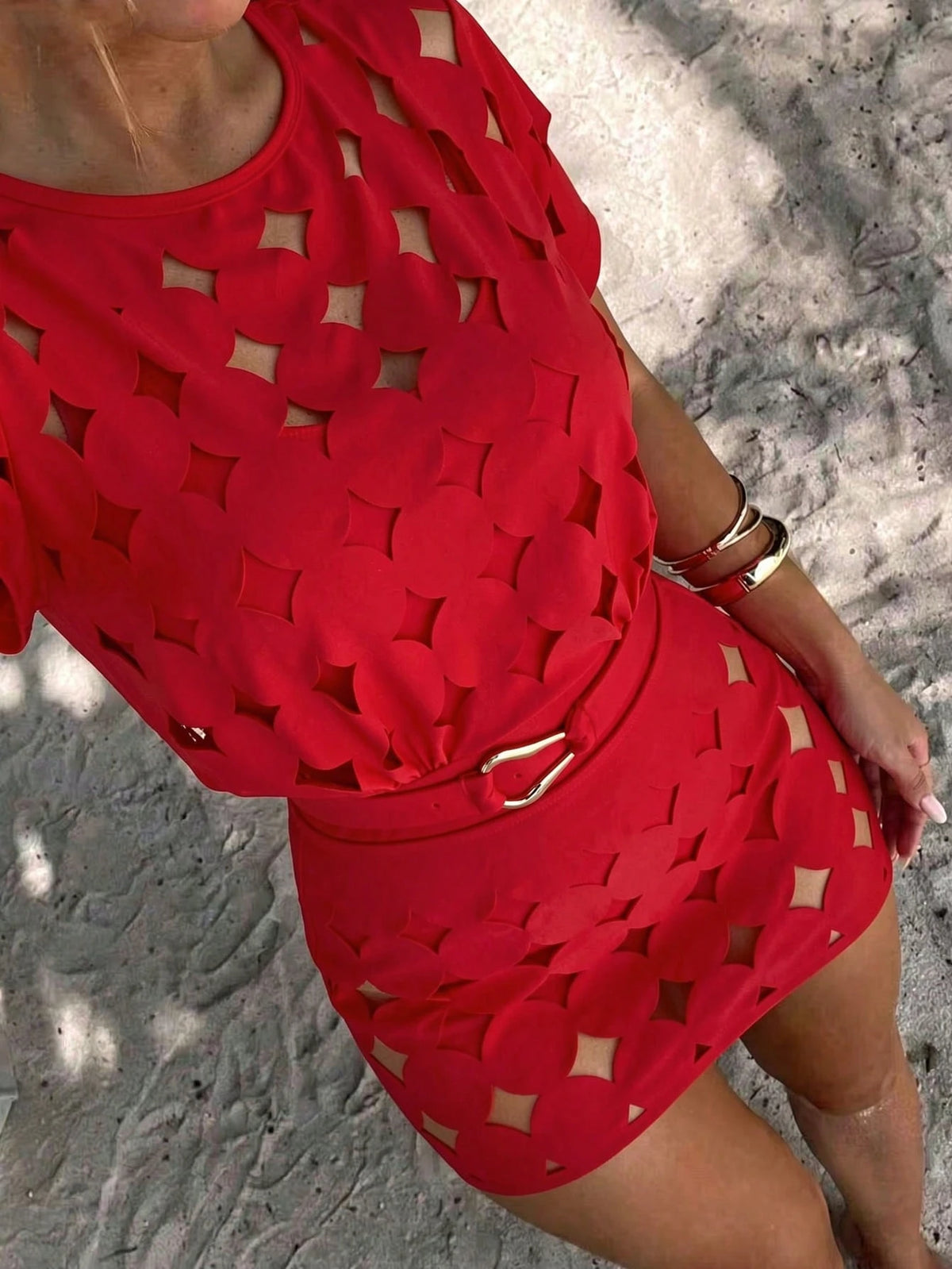 A close-up of a person wearing a bright red cutout dress with a matching belt and gold-tone buckle, standing outdoors on a textured light surface with dappled shadows.