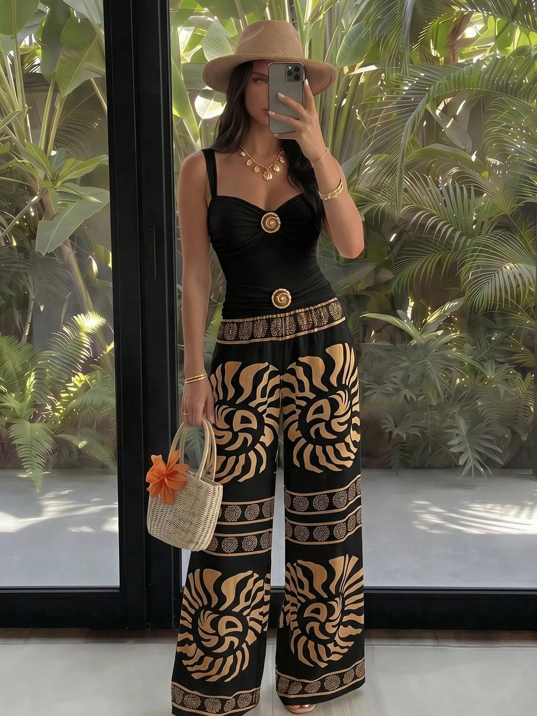 A woman takes a mirror selfie wearing a black sleeveless jumpsuit with wide legs featuring a bold tan and black zebra-like pattern, a matching belt with a gold medallion, a beige wide-brim hat, and carrying a woven straw bag with an orange flower accent.