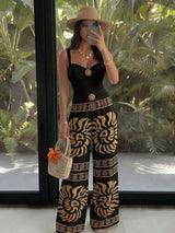 A woman takes a mirror selfie wearing a black sleeveless jumpsuit with wide legs featuring a bold tan and black zebra-like pattern, a matching belt with a gold medallion, a beige wide-brim hat, and carrying a woven straw bag with an orange flower accent.
