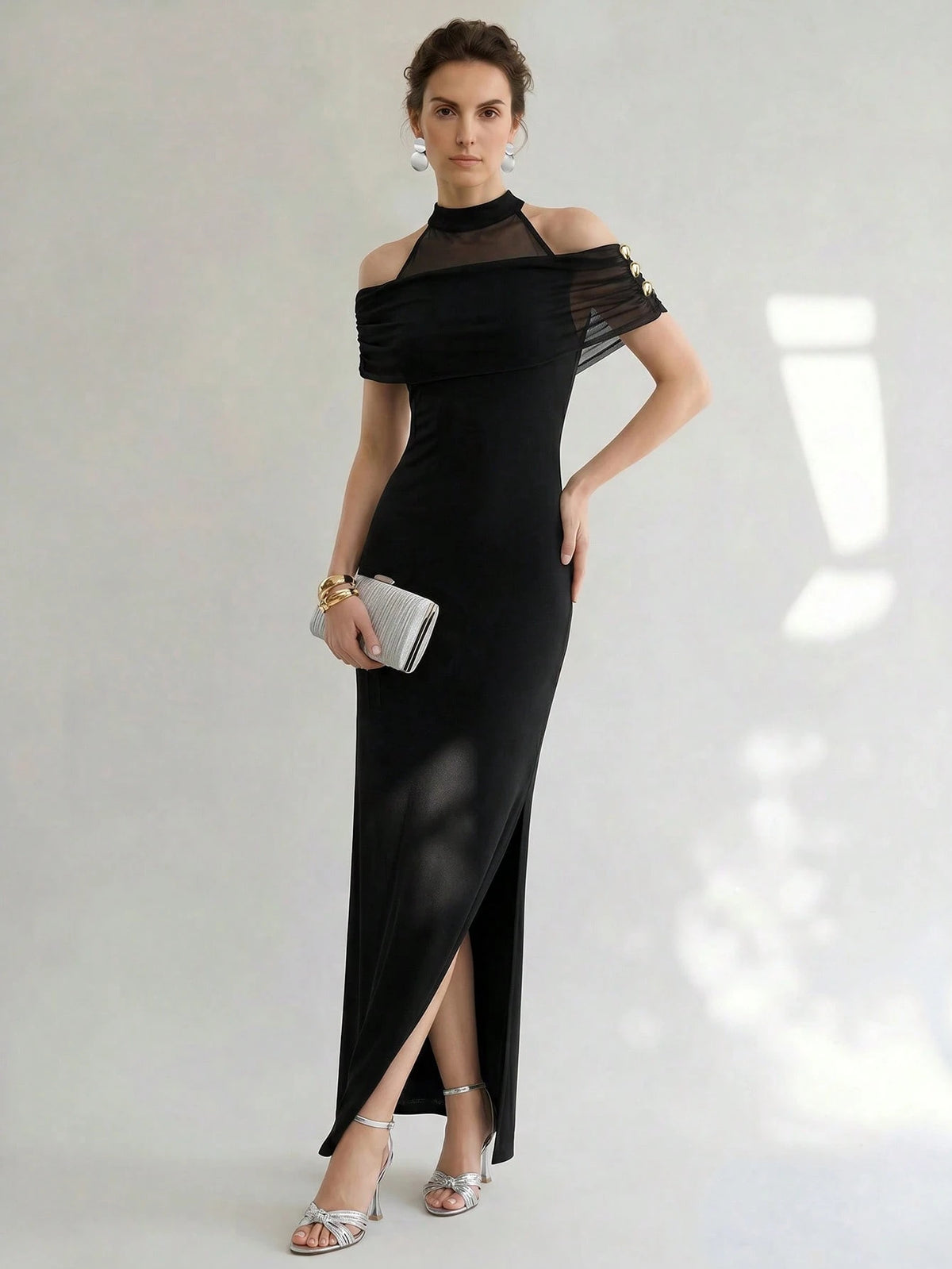 A woman models a black off-the-shoulder maxi dress with sheer mesh sleeves and a high slit, holding a small silver clutch against a light gray studio background.