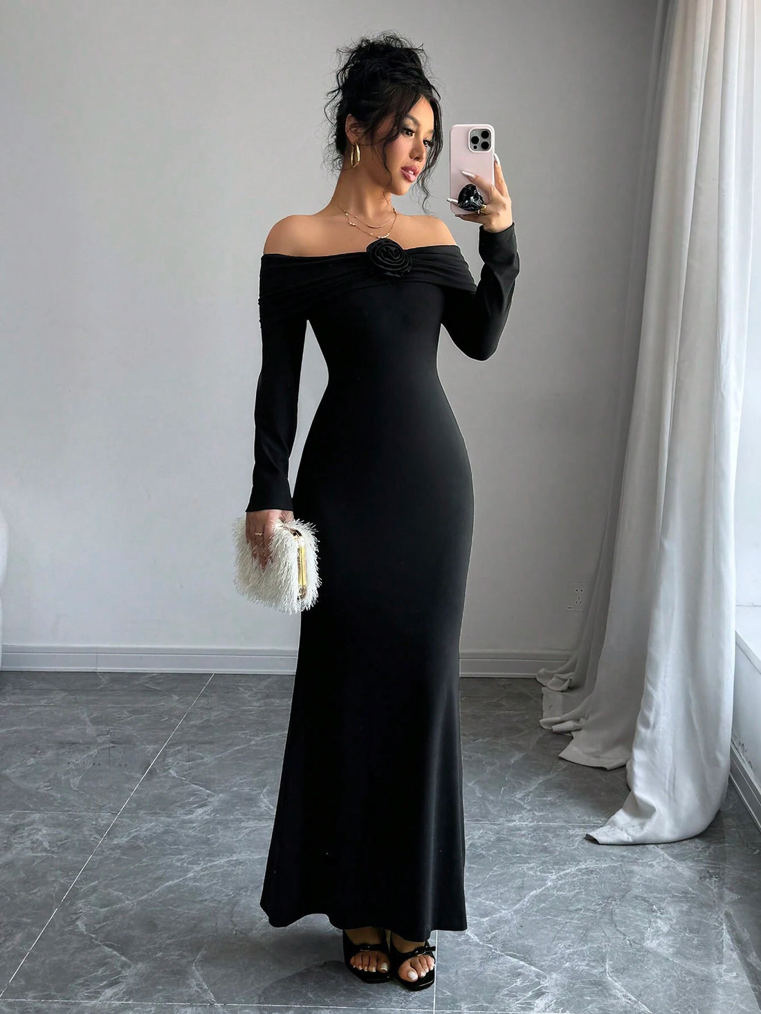 A woman takes a mirror selfie wearing a long-sleeve off-the-shoulder black maxi dress with a ruched detail at the neckline. She is holding a small white textured clutch and wearing black strappy heels, standing in a room with gray tiled floors and a white curtain.