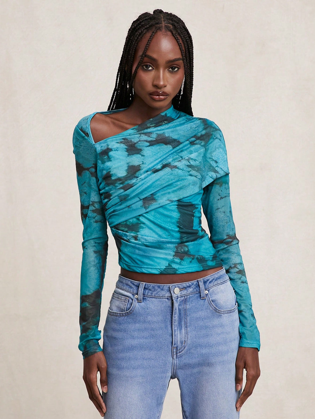 A woman modeling a long-sleeve asymmetrical wrap top with a teal and black tie-dye pattern, paired with light-wash blue jeans, against a neutral beige background.