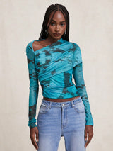 A woman modeling a long-sleeve asymmetrical wrap top with a teal and black tie-dye pattern, paired with light-wash blue jeans, against a neutral beige background.