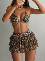 A woman modeling a two-piece leopard print swimsuit consisting of a halter triangle bikini top and a ruffled high-waisted mini skirt, set against a plain light gray background.